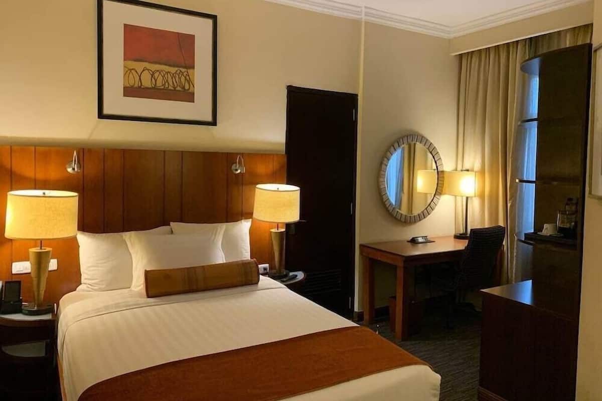 A hotel room with a bed, two bedside lamps, a desk with a chair, a mirror, and a framed picture on the wall.