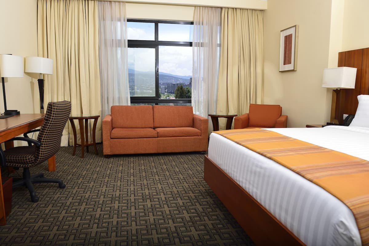 A hotel room with a bed, two chairs, a desk, and a view of a cityscape through a large window.