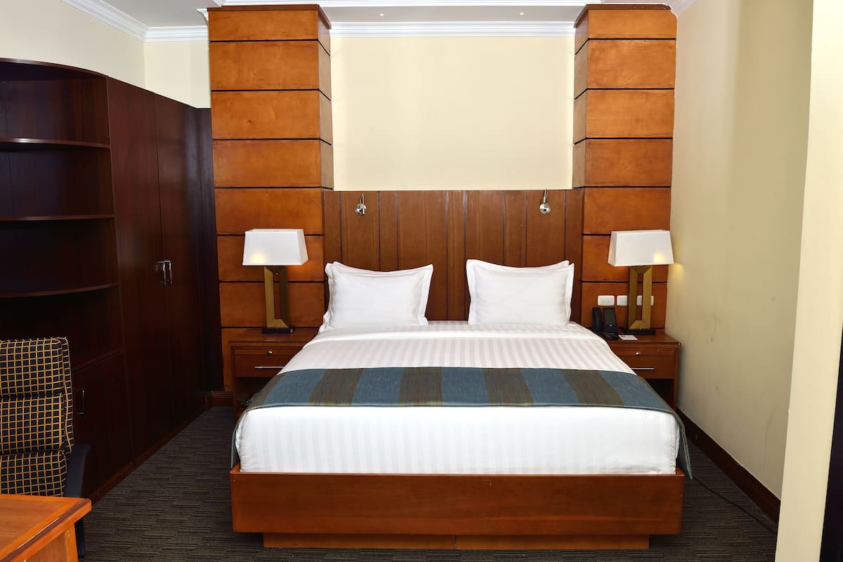 A hotel room with a large bed, wooden headboard, two bedside tables with lamps, a wardrobe, and a seating area.