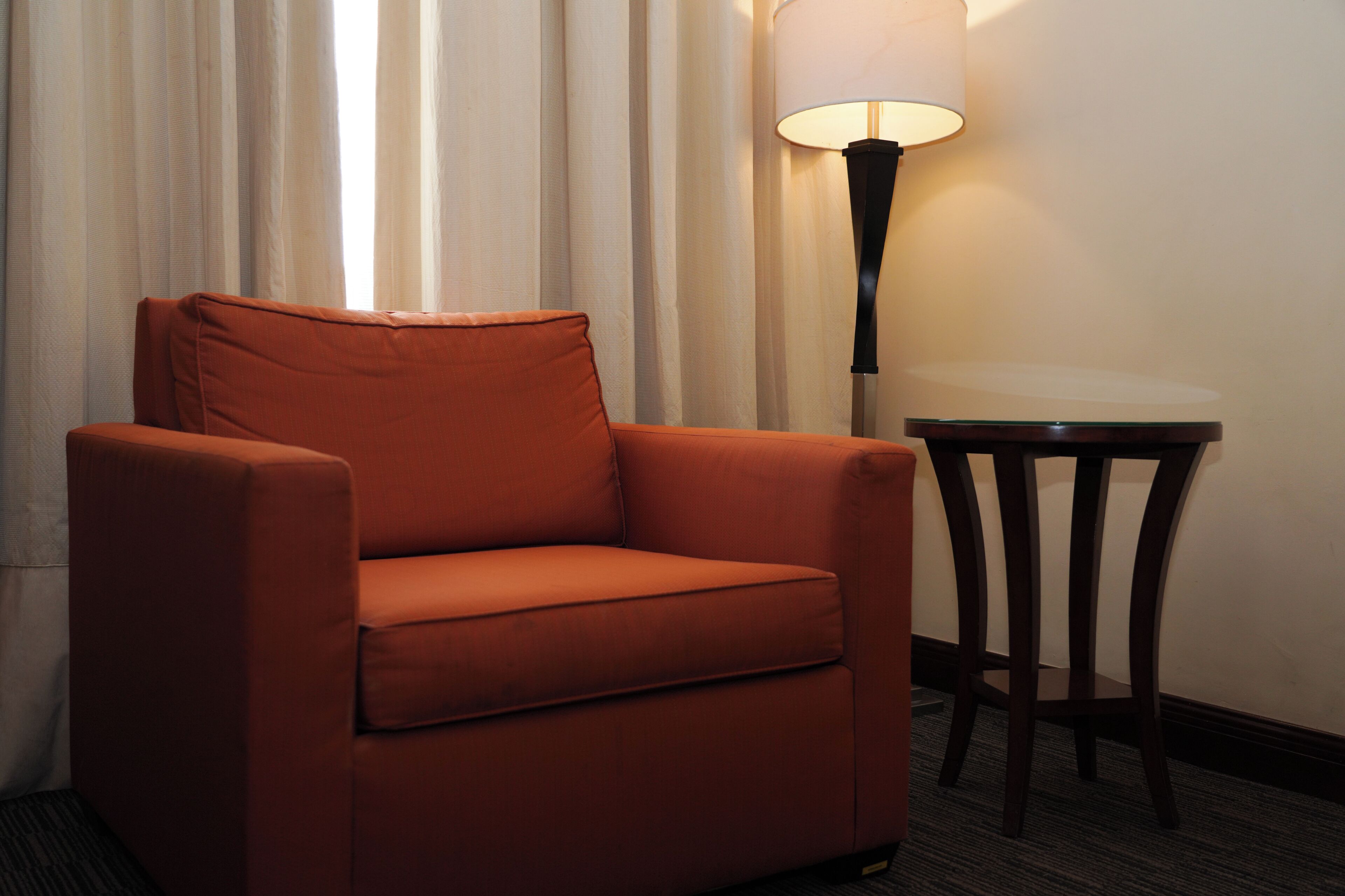 Un sillón naranja solitario en una esquina, con una pequeña mesa redonda a su lado, una lámpara sobre la mesa y una cortina al fondo.