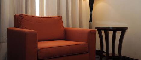 Un sillón naranja solitario en una esquina, con una pequeña mesa redonda a su lado, una lámpara sobre la mesa y una cortina al fondo.