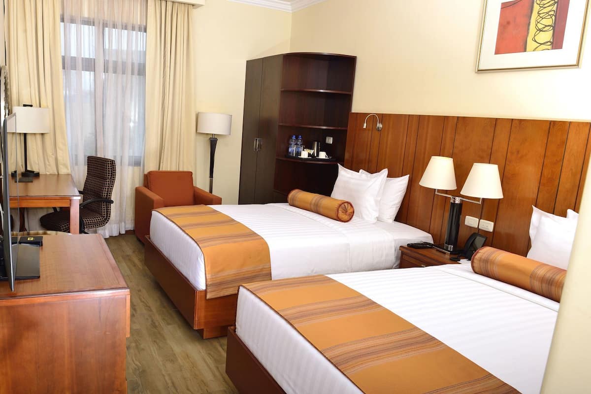 A hotel room with two beds, a desk, a chair, a wardrobe, and a lamp.