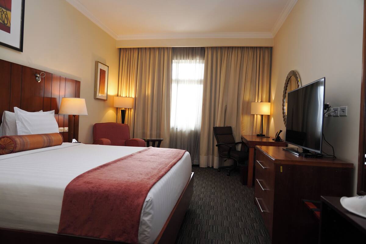 A hotel room with a large bed, a desk with a TV, a chair, and a desk lamp.
