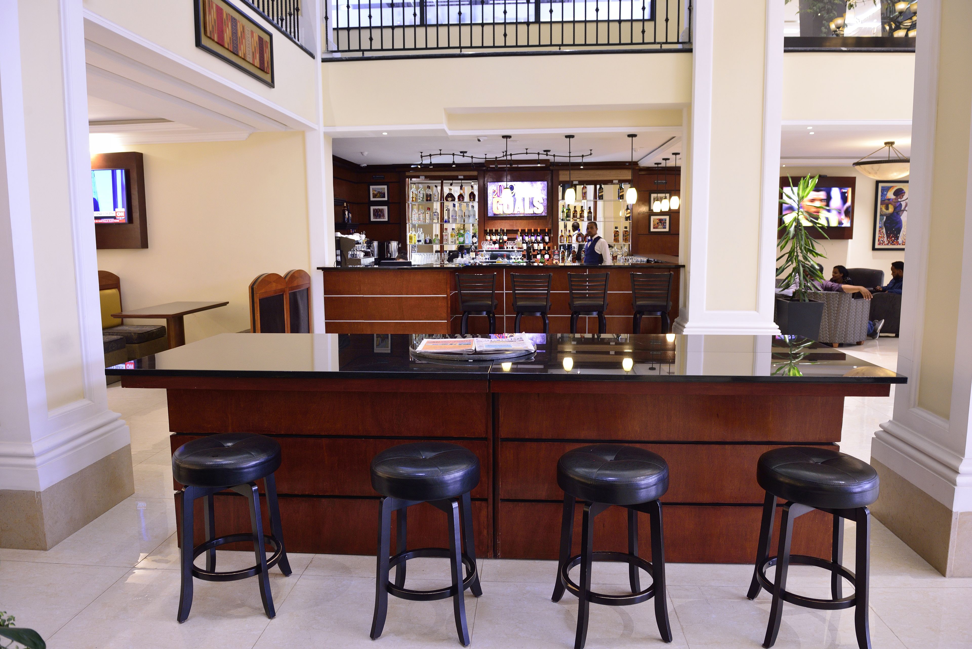 A spacious bar area with a large counter and three stools, a television mounted on the wall, and a variety of bottles and glasses on shelves.
