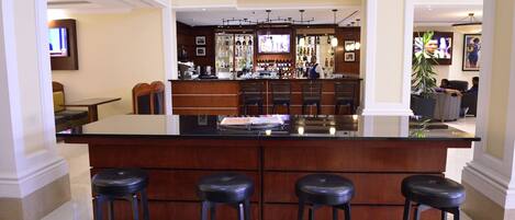 A spacious bar area with a large counter and three stools, a television mounted on the wall, and a variety of bottles and glasses on shelves.