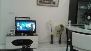 Sala de estar con televisor de pantalla plana sobre un soporte blanco, un florero con flores y un cuadro en la pared.