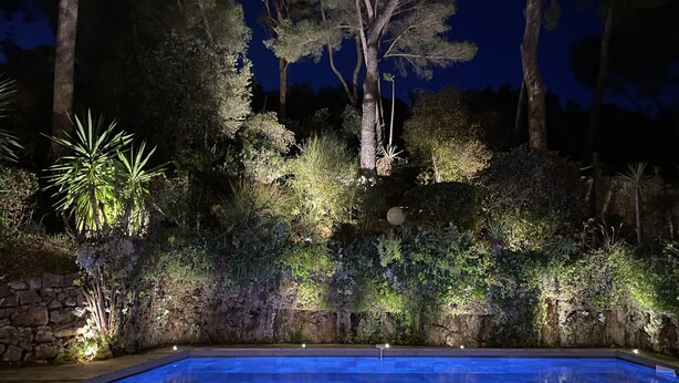 Une piscine dotée d’une terrasse en bois, entourée d’une végétation luxuriante et de murs en pierre, le tout illuminé la nuit.