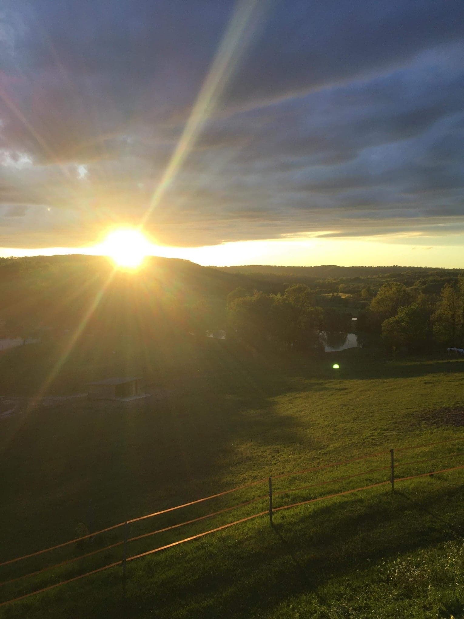 Ein Sonnenuntergang über einer hügeligen Landschaft, mit einem eingezäunten Feld und einem kleinen Gebäude in der Ferne.