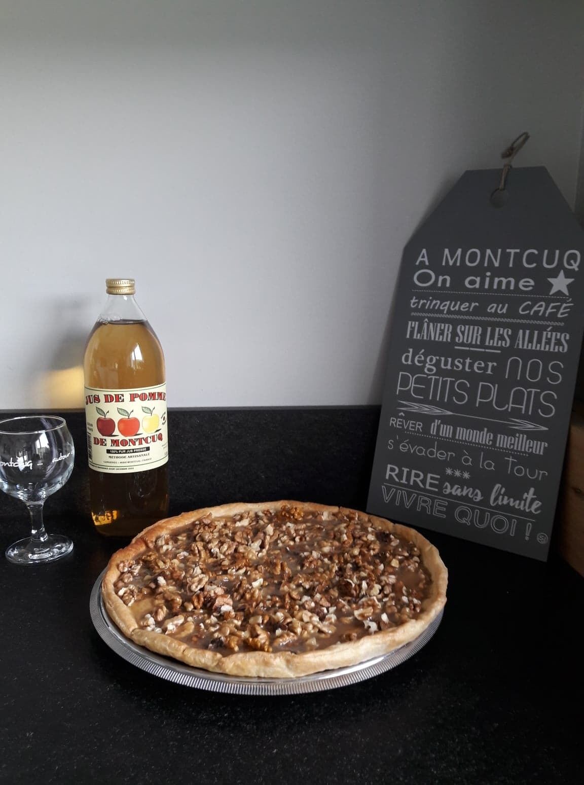 Una botella de vino Montcuq, una pizza y un cartel con texto en francés sobre una encimera de cocina.