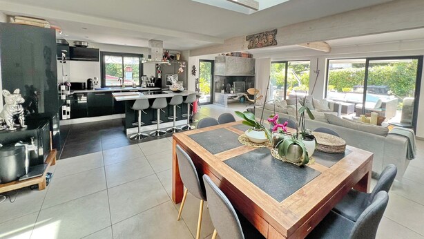 A modern kitchen with a central island, a dining area with a wooden table and chairs, and a living room with a sofa and large windows.