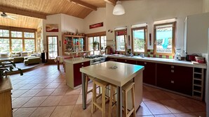 A spacious kitchen with a central island, wooden ceiling beams, large windows, and a dining area with a sofa and table.