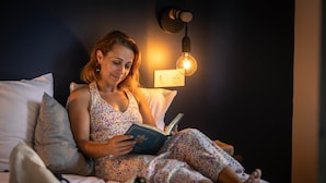 Une personne est assise sur un lit, en train de lire un livre dans une pièce où une lampe est fixée au mur.
