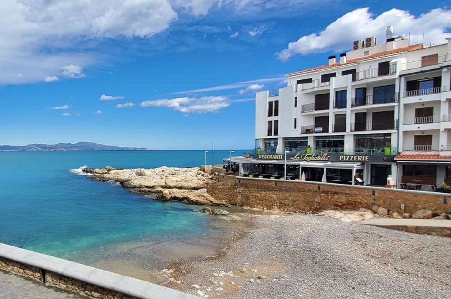 A coastal building with a restaurant and pizzeria on the ground floor, overlooking a rocky shoreline and clear blue sea.