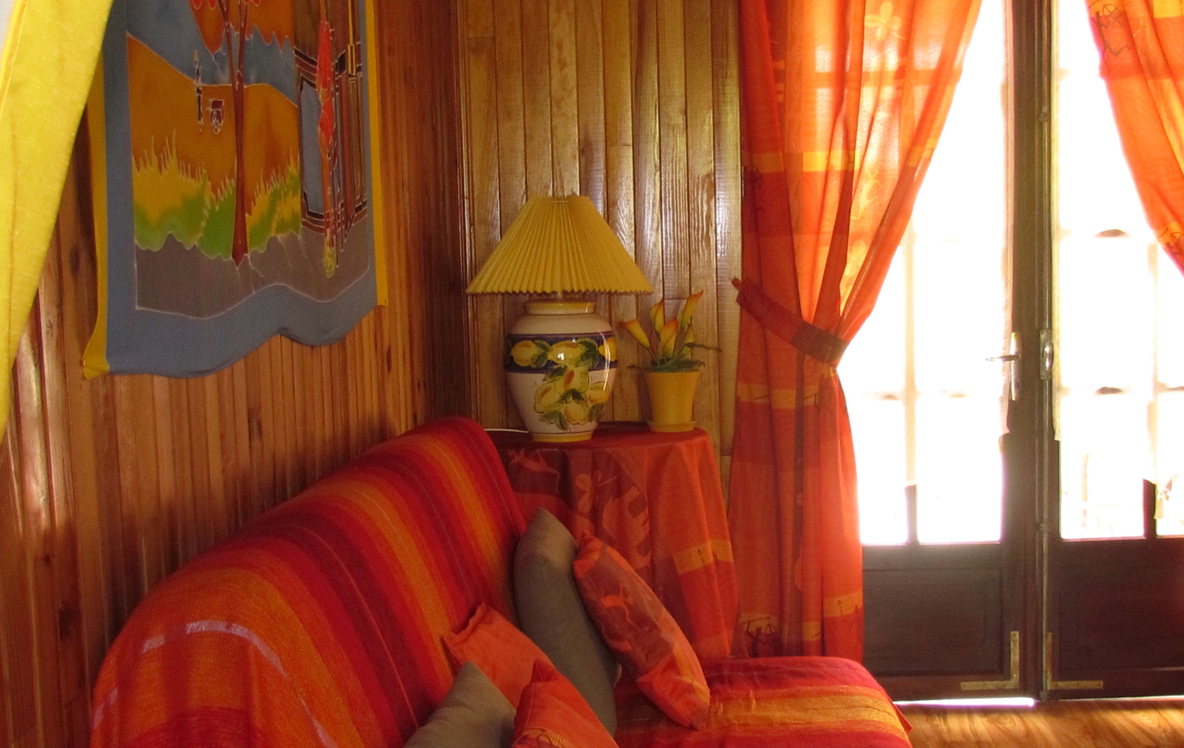 Quarto com parede de madeira, sofá com almofadas laranja, luminária e janela com cortinas.