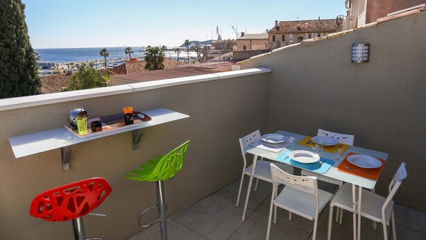 A balcony with a table set for dining, white chairs, and a view of the beach and buildings. - DUPLEX WITH GARAGE BETWEEN BEACHES AND TOWN CENTER (La Ciotat)