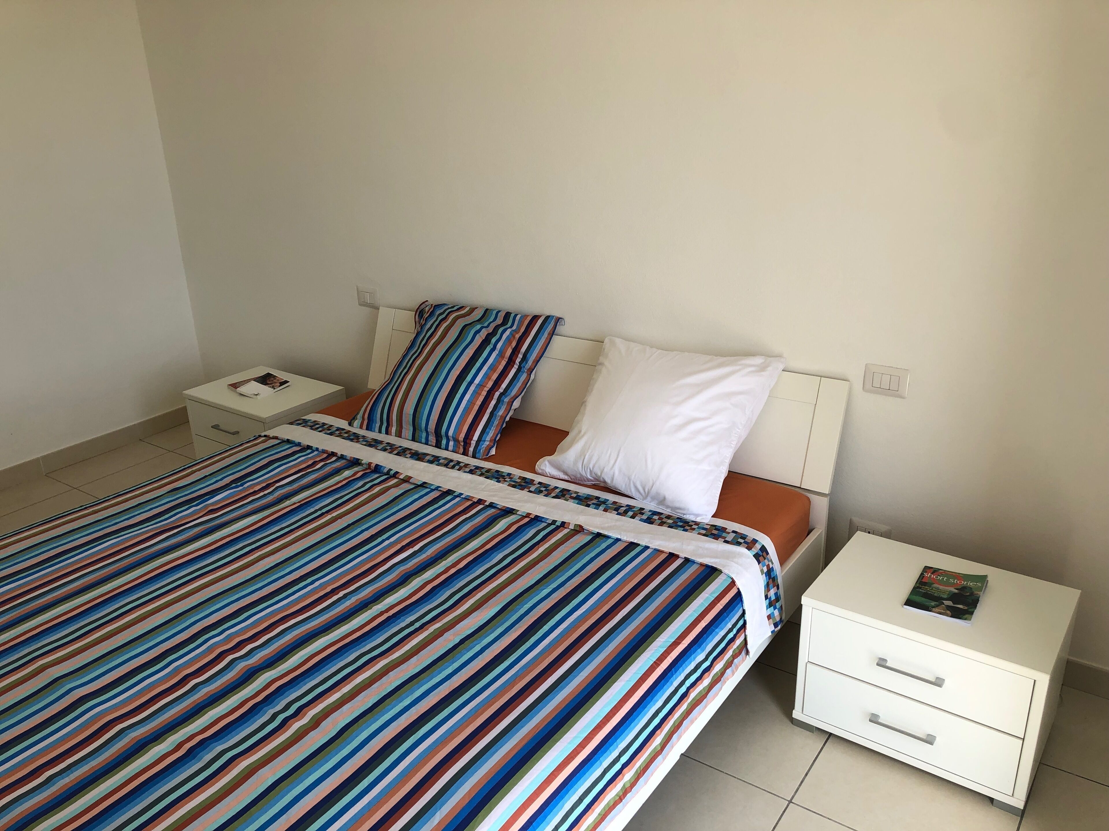 A neatly made bed with striped bedding, a white pillow, and a white nightstand with drawers.
