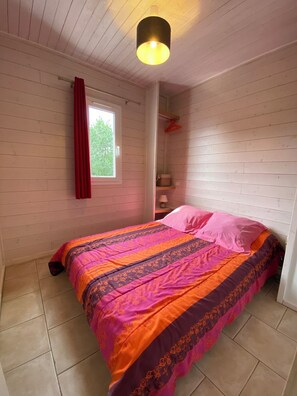 Cozy bedroom with a comfortable bed against a wall, featuring a window, ceiling light, and shelf with personal touches.