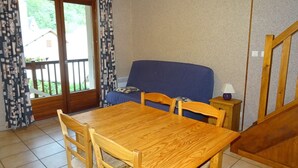 Une pièce comprenant une table et des chaises de salle à manger en bois, un canapé bleu, une petite table de chevet en bois avec une lampe, et une porte-fenêtre coulissante donnant sur un espace extérieur.