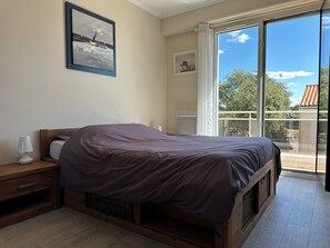 A bedroom with a bed, a nightstand, a lamp, a window with a view of trees, and a painting on the wall.
