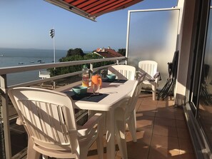 A balcony with a table set for two, a bottle of juice, a glass, and a bowl. The balcony overlooks a body of water with boats and houses in the background.
