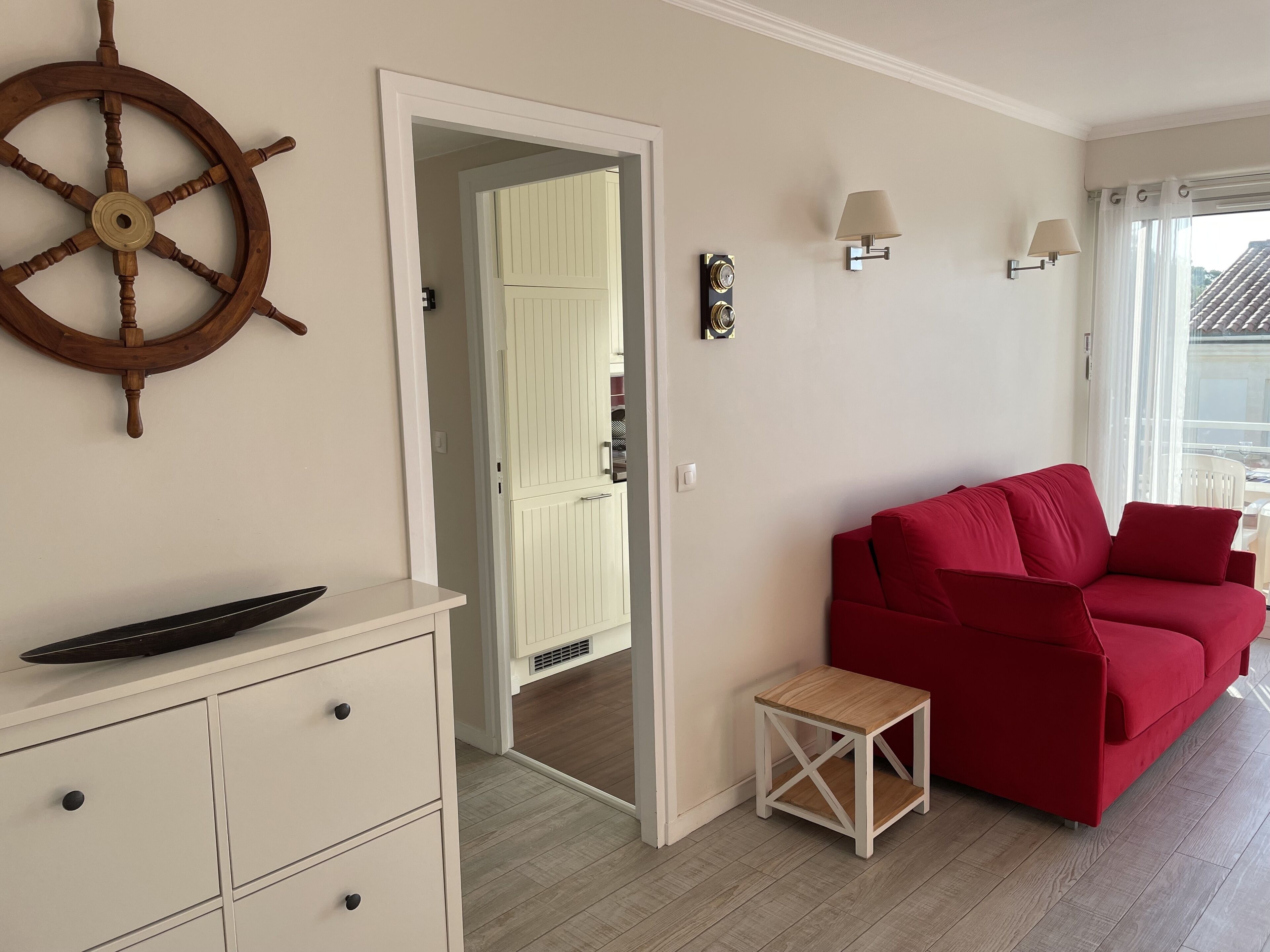 A room with a red sofa, a small wooden side table, a white dresser, a wall-mounted ship's wheel, and a door leading to another room.