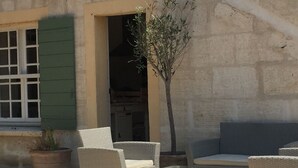 An outdoor seating area with wicker furniture and a glass table, adjacent to a building with a stone facade and a window with green shutters. - Magnificent in the heart of Provence (Maillane)