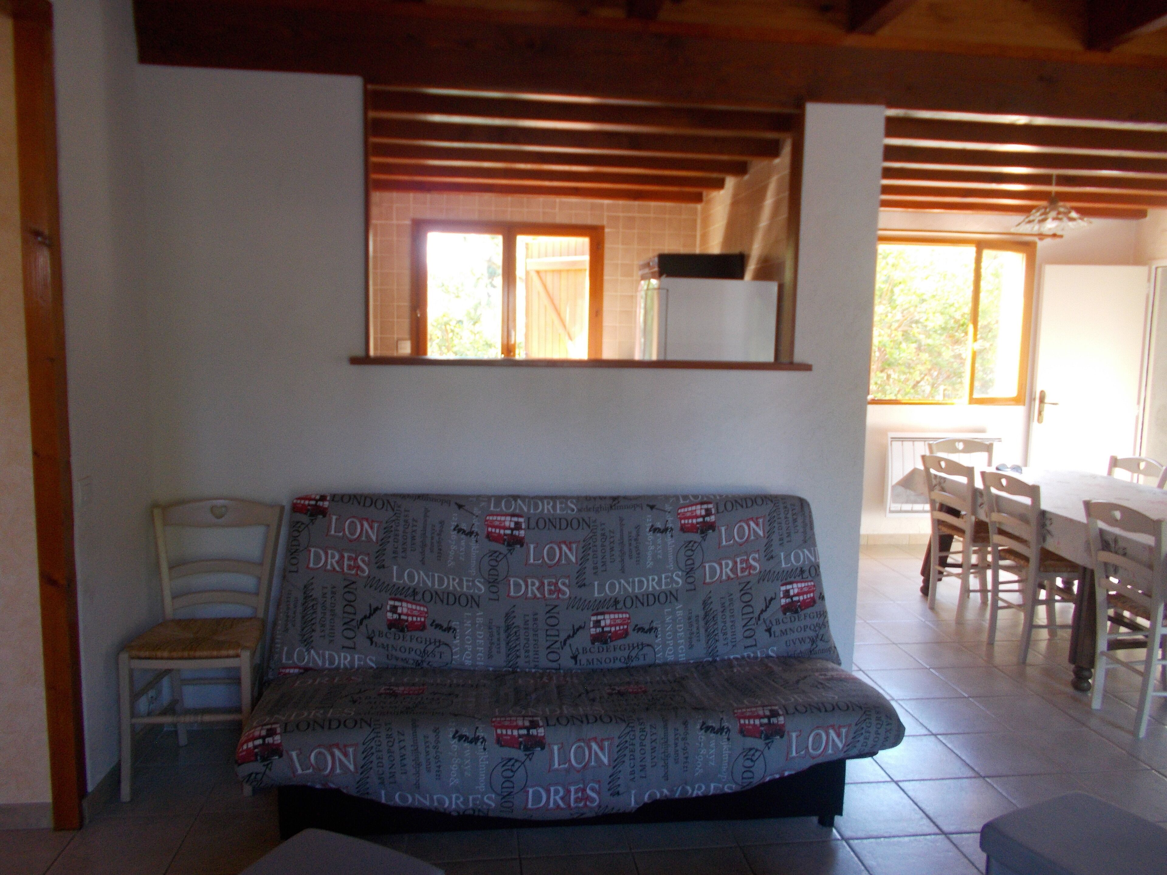 Un salon avec un canapé recouvert d’une couverture aux motifs londoniens, une chaise en bois, une table à manger avec des chaises et une fenêtre donnant sur un paysage verdoyant.