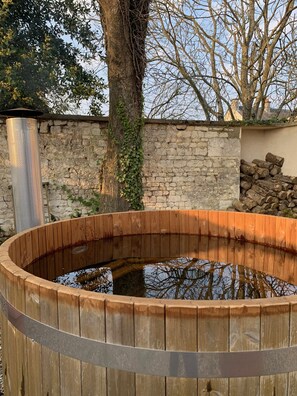 A wooden hot tub with a stone wall and a tree in the background.