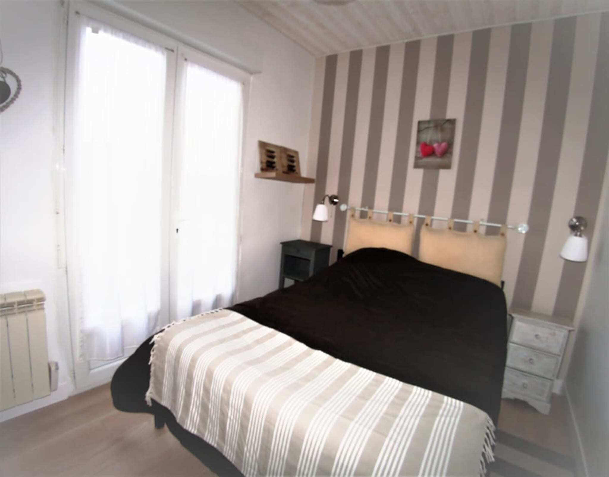 Een slaapkamer met een bed, nachtkastjes, een radiator, een raam en gestreept behang.