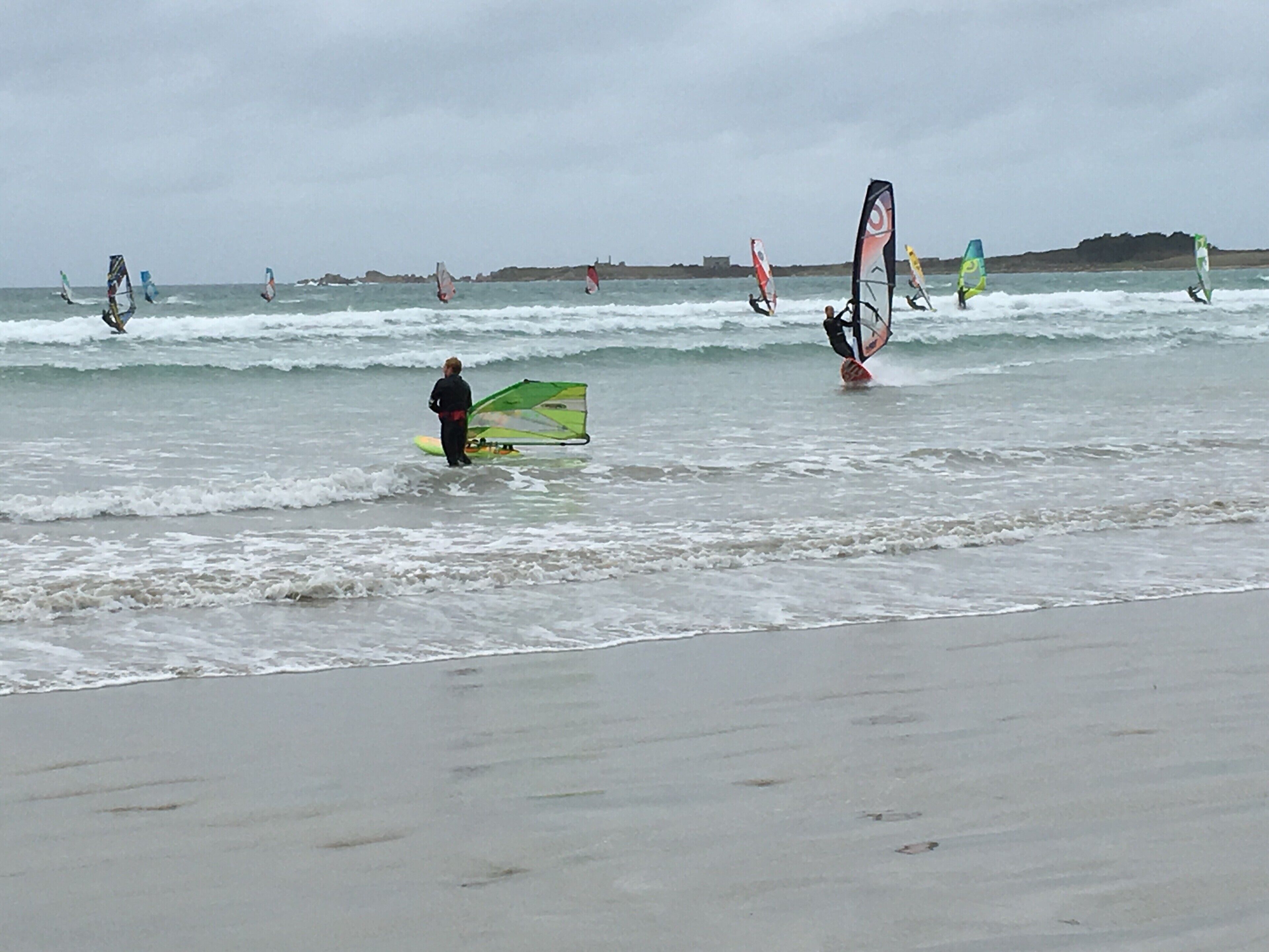 A beach with multiple windsurfers in the water and one standing on the shore with a windsurf board.