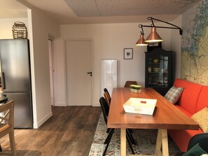 A modern kitchen with a refrigerator, wooden dining table, and chairs. There is a red sofa and a cabinet with glass doors.