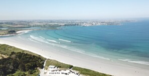An aerial view of a coastal beach with a parking area and buildings in the distance.