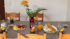 Une table à manger est dressée avec une variét é d’aliments, notamment des fruits, du pain et une carafe de jus d’orange. On y trouve également plusieurs verres à vin et un vase rempli de fleurs.