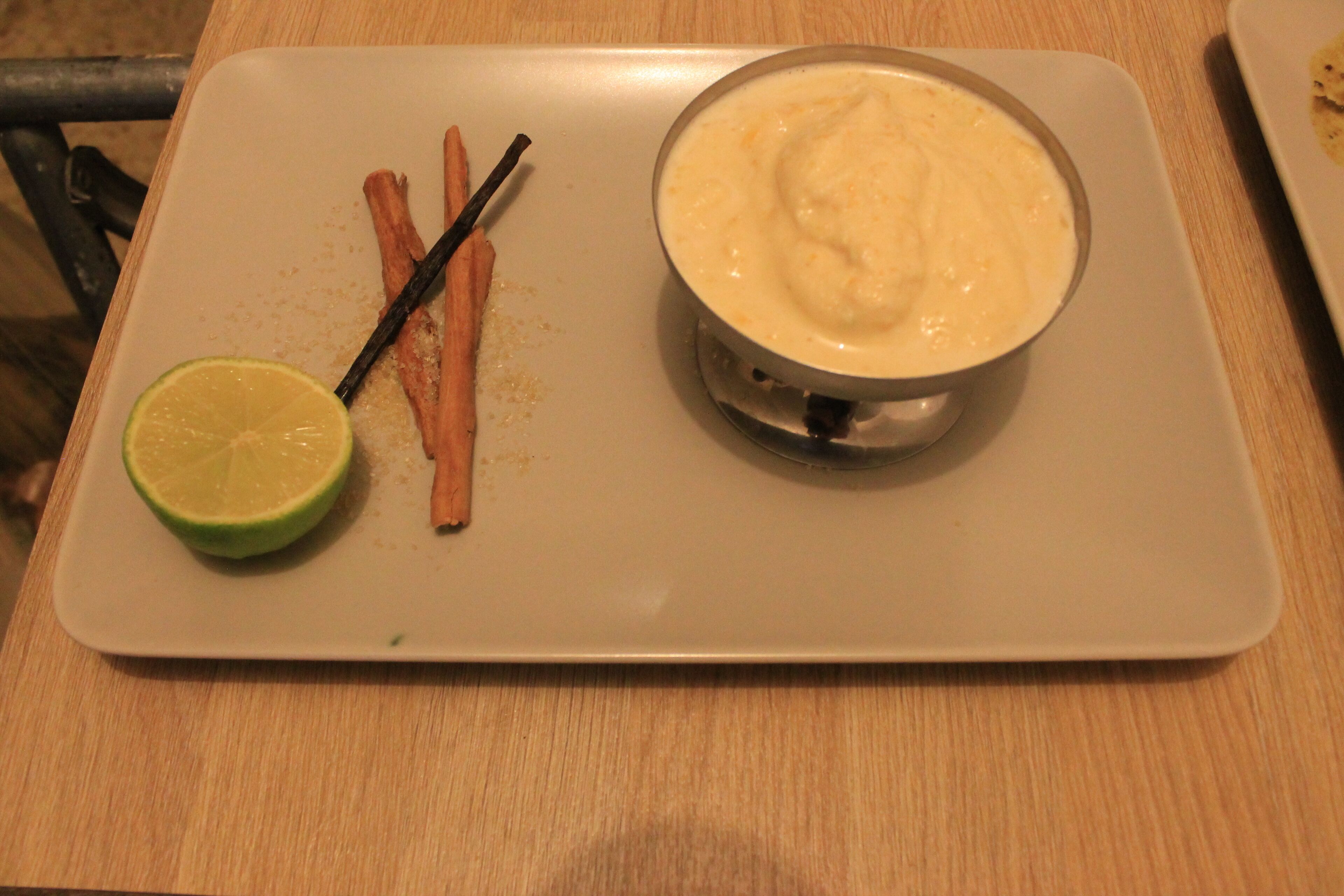 A white plate with a bowl of creamy dip, a lime wedge, and two sticks of cinnamon on a wooden table.