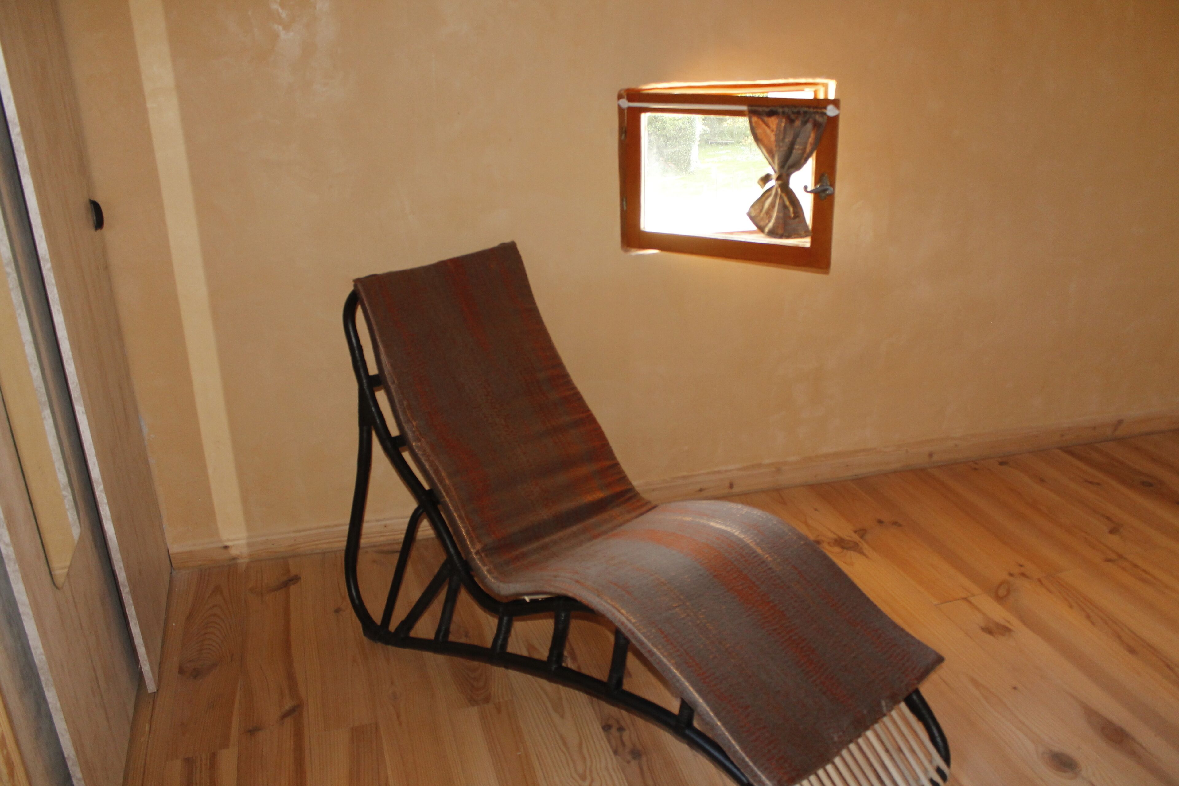 A room with a wooden floor, a single brown chair, and a small window with curtains.