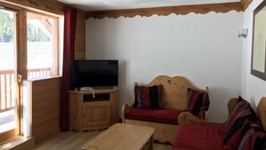 Een kamer met een houten vloer, een televisie op een houten standaard, een houten bank met rode kussens en een houten tafel.