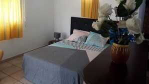 Ein Schlafzimmer mit einem Bett, einem Nachttisch, einer Lampe, einer Vase mit Blumen und Vorhängen.