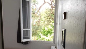A small, compact room with a single bed, a chair, a table, a window with curtains, and a radiator.
