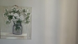 A room with a table featuring a geometric patterned tablecloth, a vase with white flowers, and a painting of a vase with flowers on the wall.