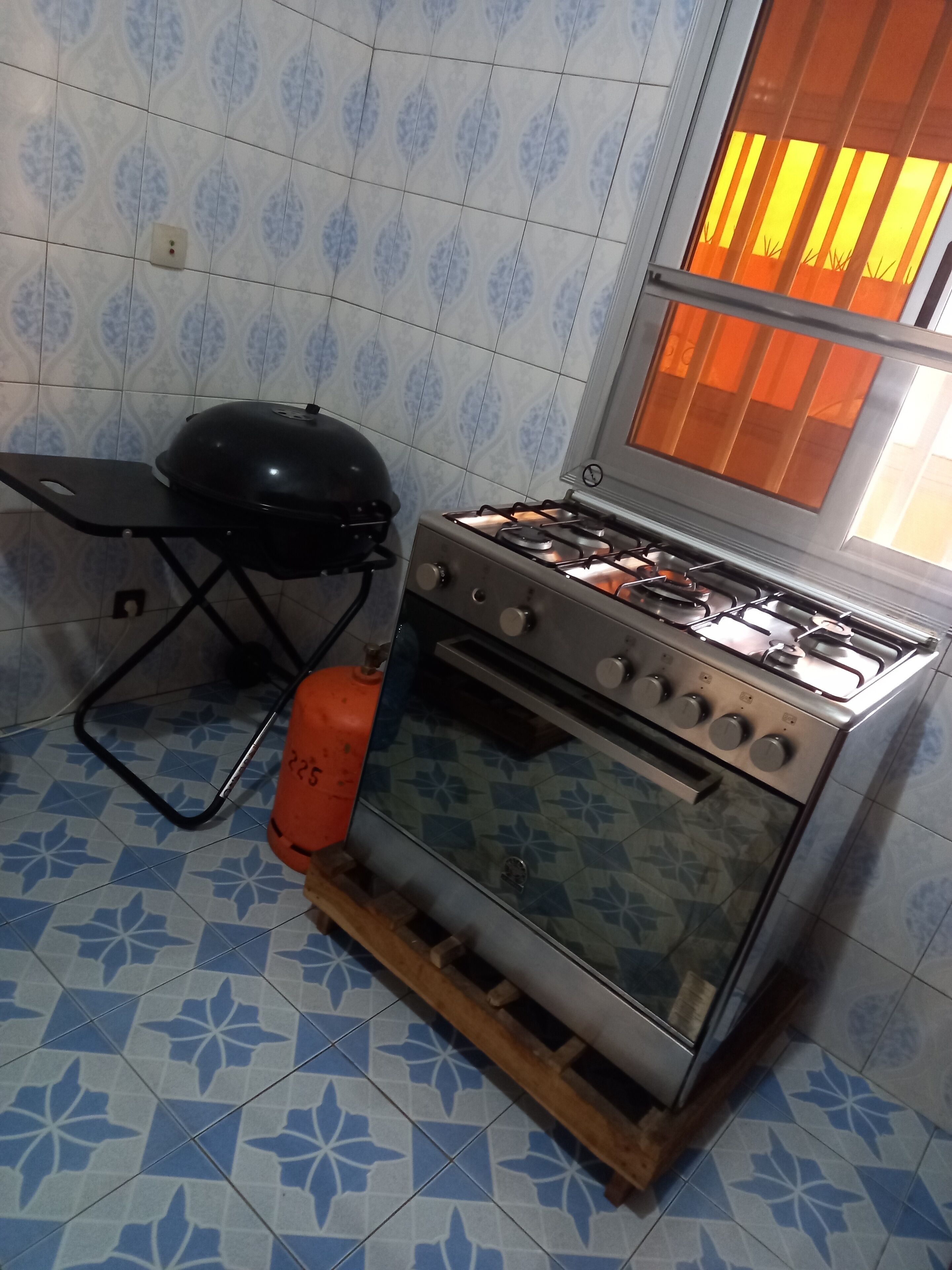 Una cocina con estufa a gas, una parrilla portátil y un ventanal con cortinas anaranjadas.