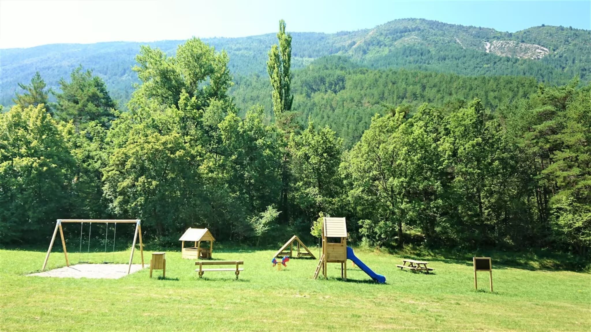 Un parque infantil con columpios, un tobogán y una casita en un campo cubierto de césped, con una montaña boscosa al fondo.