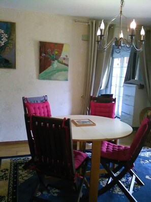 Un comedor con una mesa redonda, dos sillas, una lámpara de araña, un cuadro en la pared y una ventana con cortinas.