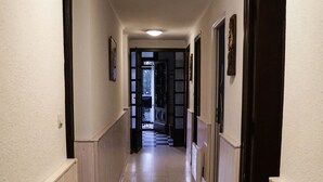 Um corredor estreito, paredes brancas e chão em mosaico, conduzindo a uma porta no final.