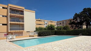 A swimming pool with stone tiles, a building with balconies, and a hedge.