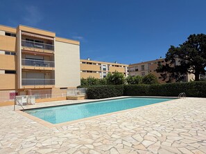 A swimming pool with stone tiles, a building with balconies, and a hedge.