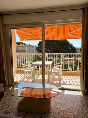 A balcony with a glass table and chairs, an orange striped umbrella, and a view of buildings and trees.