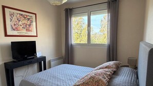 A bedroom with a bed, a TV, a desk, a radiator, and a window with curtains.