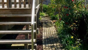 Una scala in legno conduce a un edificio con tetto in mattoni, immerso in un giardino ricco di piante e fiori.
