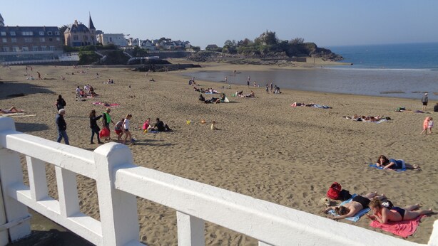Una spiaggia con persone che prendono il sole e nuotano, una recinzione bianca e edifici sullo sfondo.