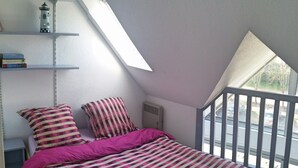Una camera da letto con soffitto spiovente, un letto con copripiumino a righe, una finestra vista strada e uno scaffale con libri e un oggetto decorativo.